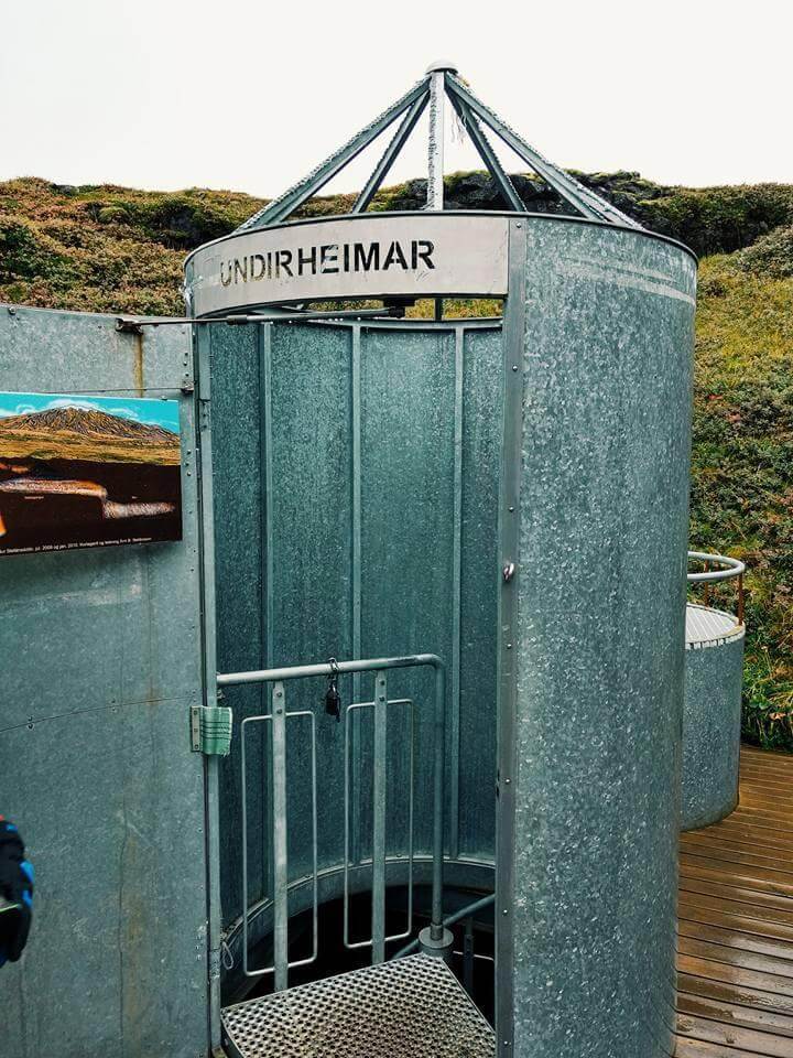 Picture of the entrance to the cave that may lead to the center of the Earth. It's a tall metal cylindrical entrance with steps leading down into the cave. The word Undirheimar stencilled across the top. The whole thing sits on a wooden platform with the rugged Icelandic terrain in the background